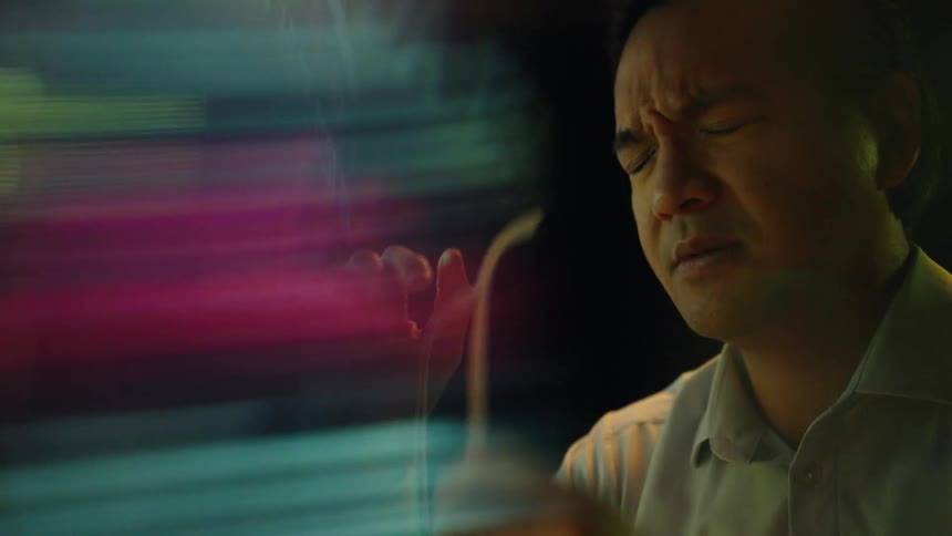 Adult man removing his glasses and pinching his nose between the eyebrows with a nervous facial expression in front of floating colorful blurred text and lights indoors