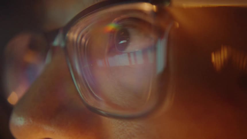 A close view of a person wearing glasses shows the reflection of a digital screen in their eyes while warm light glows in a dark room