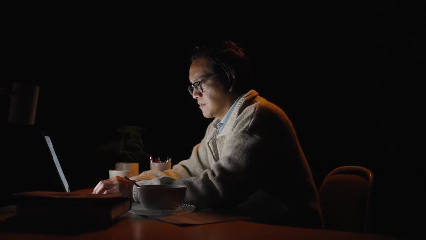 An older man wearing glasses and a warm cardigan sits at a desk in a dark room intently focused on his laptop screena small desk lamp casts a warm glow over his workspace highlighting a bowl of food and some books as he works late into th