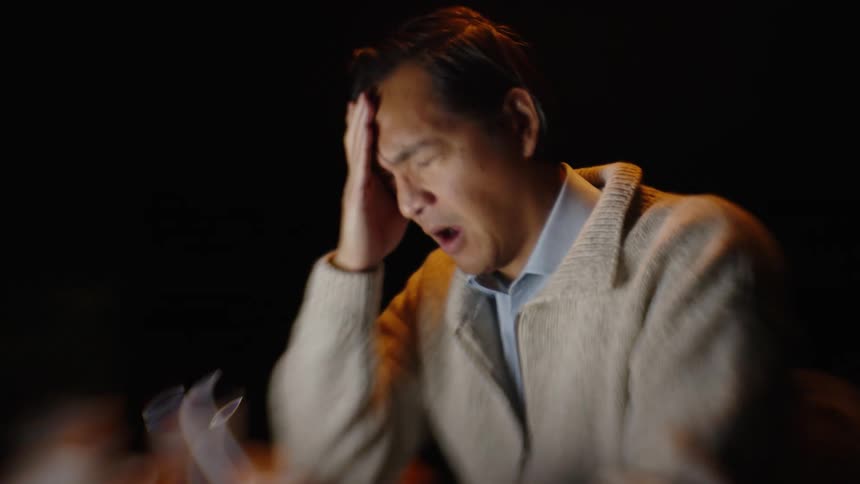 Adult man wearing a white sweater sitting down at a desk and removing his glasses then rubbing his face and yawning indoors