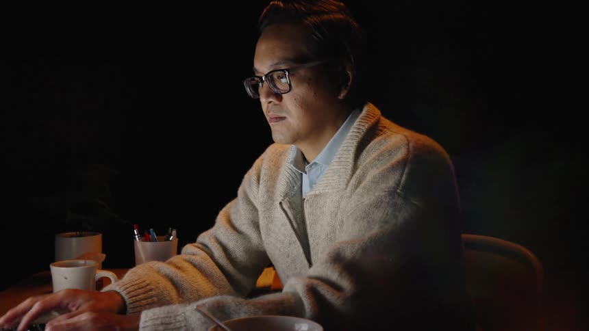 An adult man with glasses sits at a wooden desk in a dark room intently typing on a laptop while a soft warm light illuminates his face and a bowl of food nearby