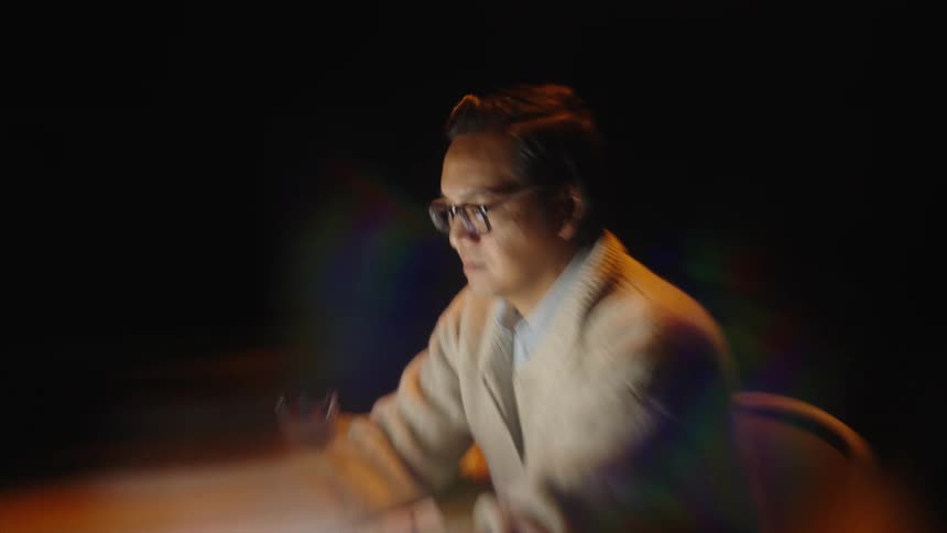 An asian man wearing glasses and a cardigan sits at a desk in a dark room working on a laptop while surrounded by papers and a mug