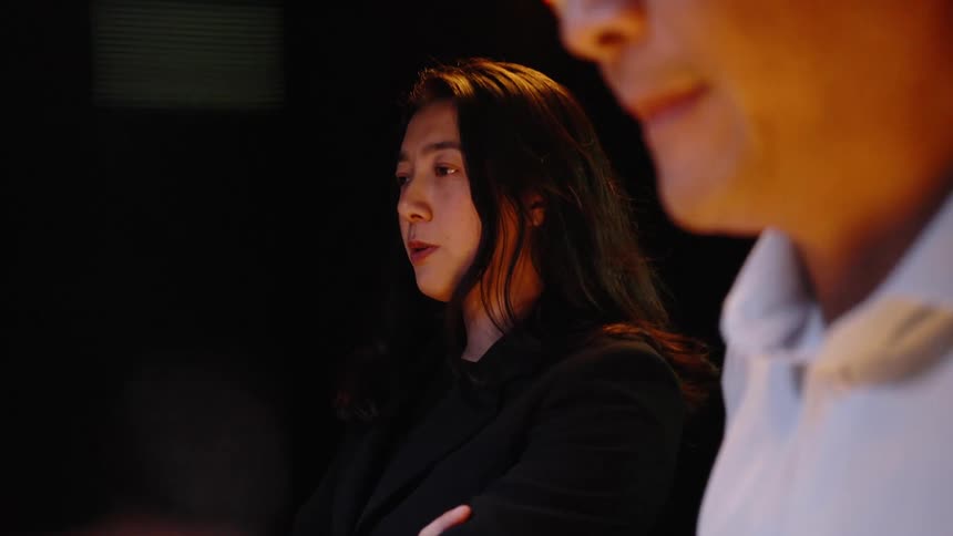 In a dark room a man and a woman in business clothes stand together while looking at a screen that provides a warm glow on their faces