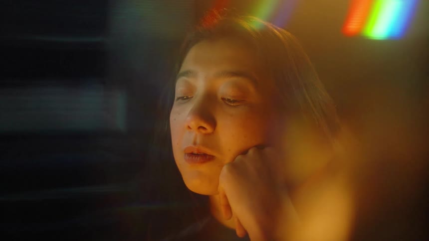 A woman wearing a black top rests her chin on her hand and looks at a screen in a dark room while soft blue light and colorful flares flicker across the foreground