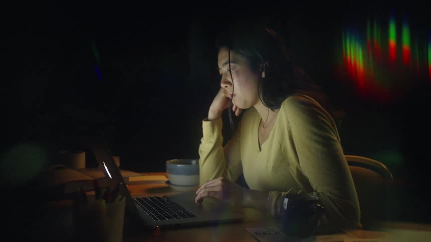 A young woman sits at a desk in a dark room her face illuminated by the glow of a laptop screen as she rests her head on her hand with a tired expressionseveral mugs and papers are scattered across the table while colorful light flares streak through