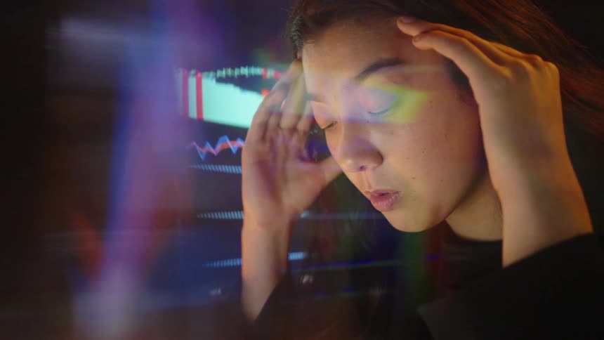 Woman holding her head in front of a monitor displaying illuminated financial charts