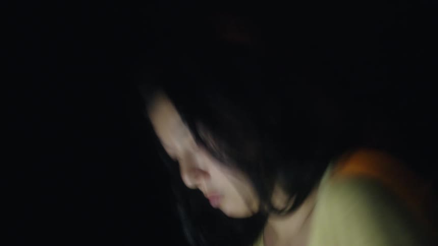A woman wearing a yellow shirt works on a laptop in a dark room with motion blur