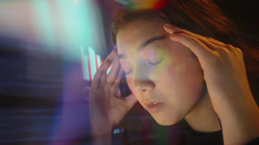 A woman wearing a black jacket sits in a dark room and presses her fingers to her temples with her eyes closed while glowing stock market charts and colorful light flares flicker in the foreground and background