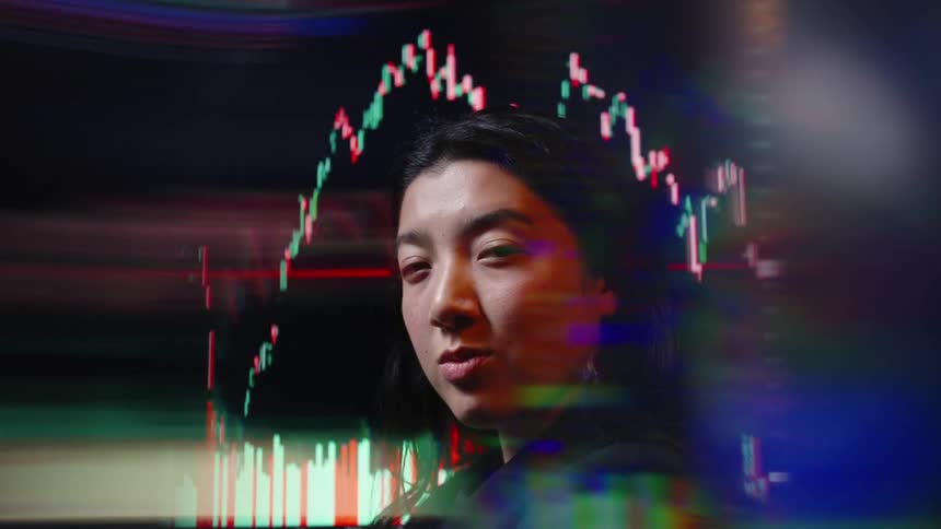 A young woman standing in front of a screen displaying stock market prices flickering behind her