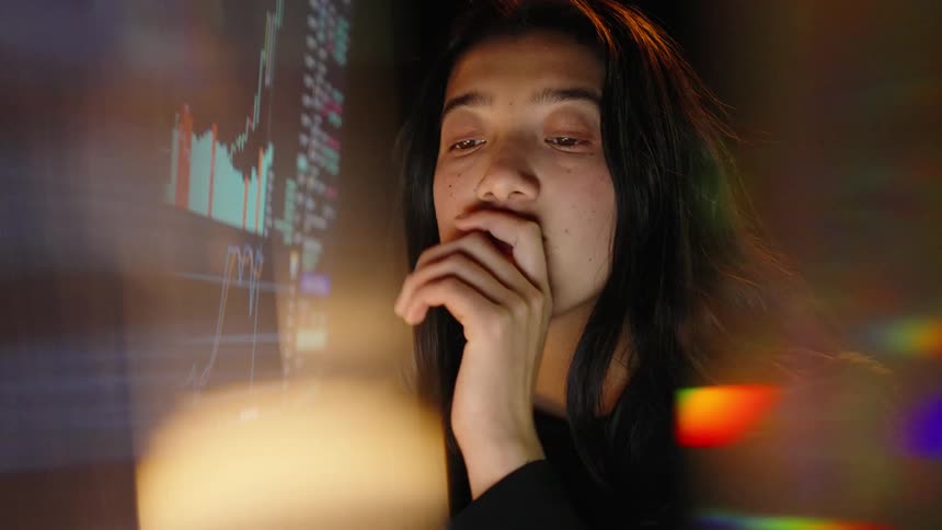 A young woman stares intently at a glowing computer screen filled with complex financial stock charts and datashe appears stressed and exhausted resting her chin on her hand before letting out a weary sigh