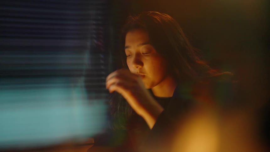 A woman wearing black clothes sits in a dark room looking at a glowing monitor with digital charts while rubbing her face and hair as warm light flares and soft blue patterns flicker in the foreground