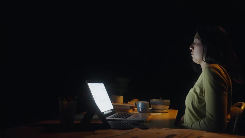 A young woman sitting at a desk behind a computer stretches her shoulders then continues typing on her pc in a dark room
