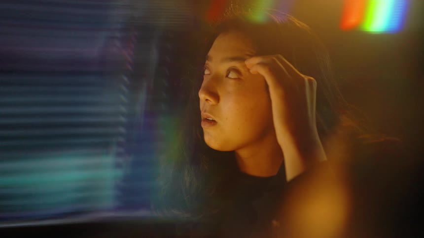 A woman wearing a black jacket sits in a dark room looking at a glowing monitor with digital stock market charts as colorful light flares and motion blur effects flicker across the foreground