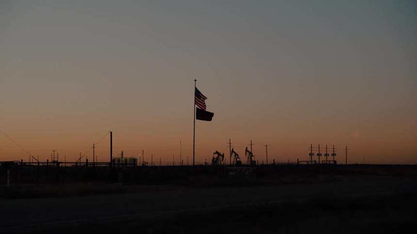 An american flag and a secondary banner flutter atop a tall pole overlooking an active oil field where pumpjacks rhythmically move against the fading glow of a wide sunset horizon