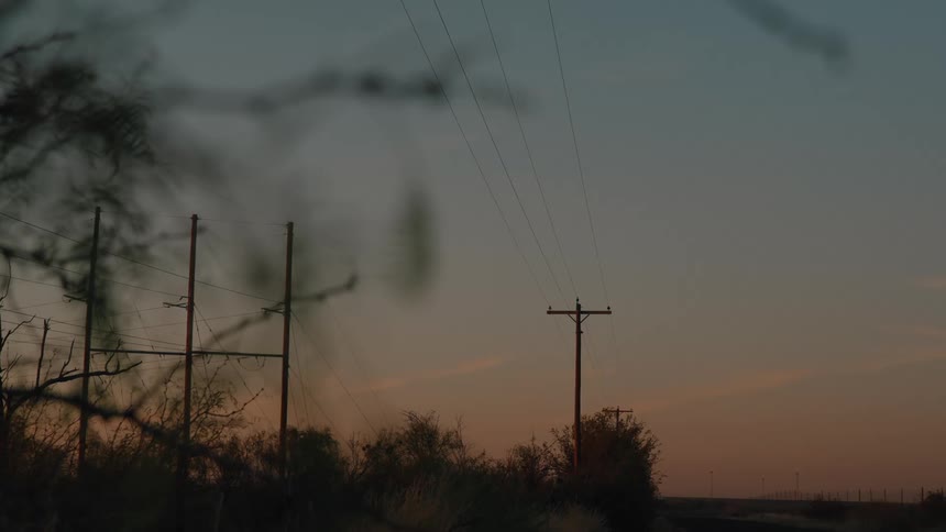 A utility pole on the side of the road holding power lines surrounded by trees at sunset