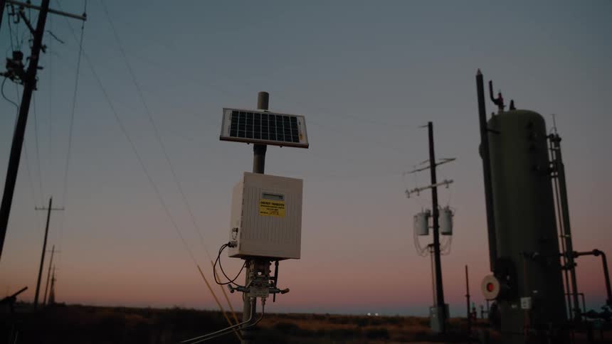 A small solarpowered device mounted on a pole stands in a rural landscape at dusk with utility poles and industrial equipment visible in the background