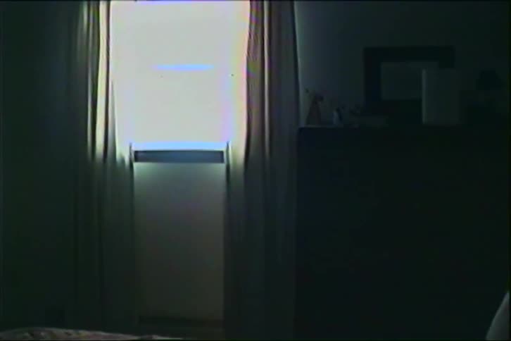 A view of a bedroom with a patterned bedspread and pillows features a small rectangular window with light filtering through thin curtains in a dark room with a grainy film texture and static distortion