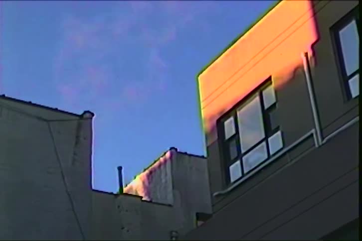 Heavy static and visual glitches continuously distort an old recording of urban buildings bathed in warm sunlight against a clear blue sky while faint steam slowly rises from a rooftop chimney