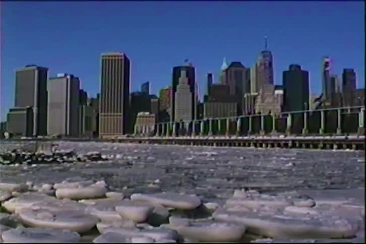 Ice floes float on the river in front of an urban skyline under a blue sky