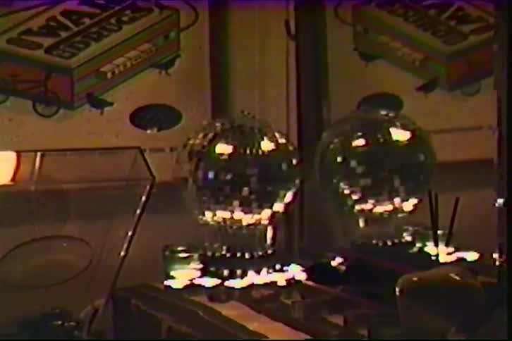 A close view of a shiny disco ball rotating in a dark room with a grainy film texture and static distortion as it reflects warm light near posters and music equipment