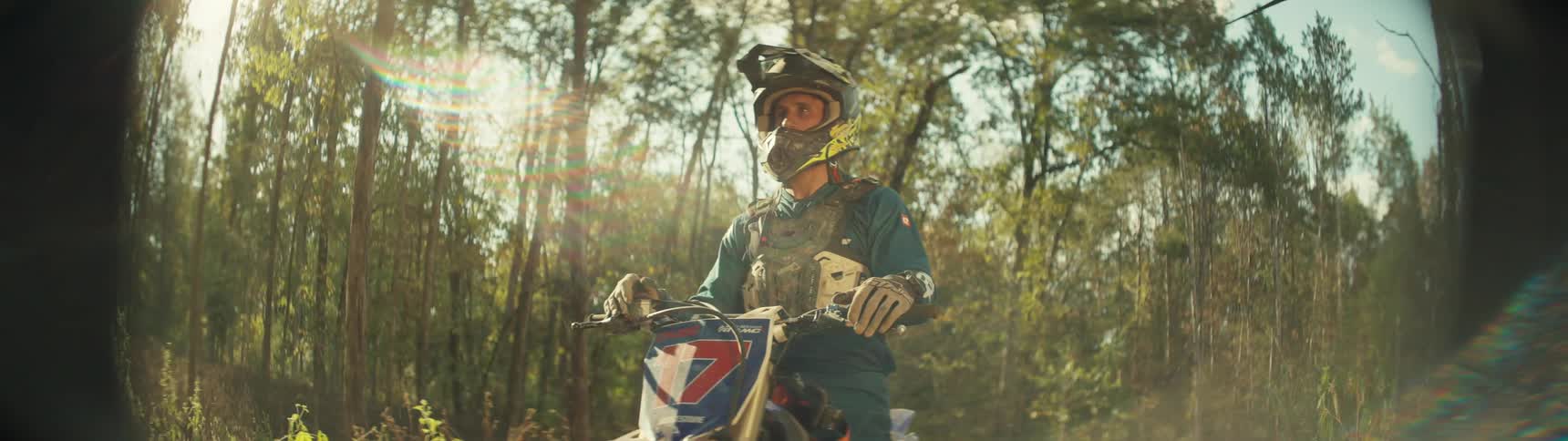 Rider wearing helmet and gloves rides a dirt bike on a trail surrounded by trees under strong sunlight