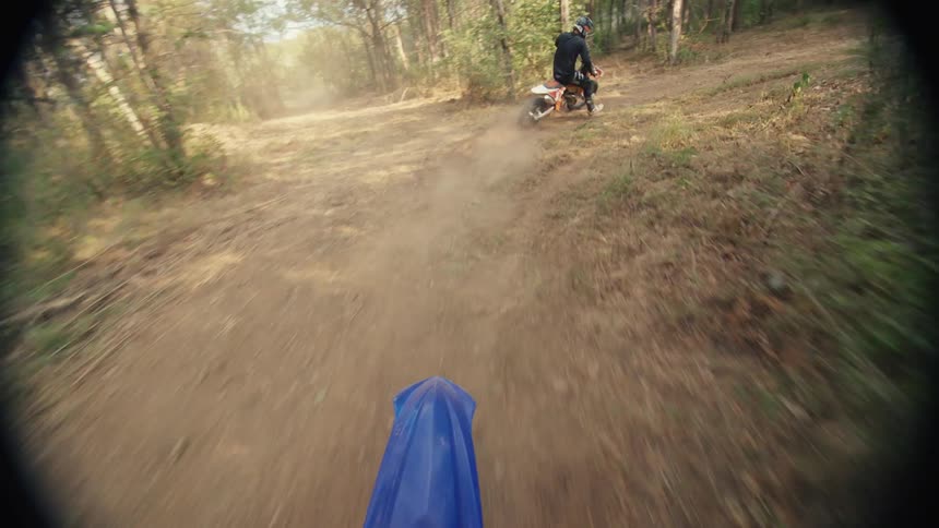A point of view shot from a dirt bike shows another rider on a dusty trail through a forest