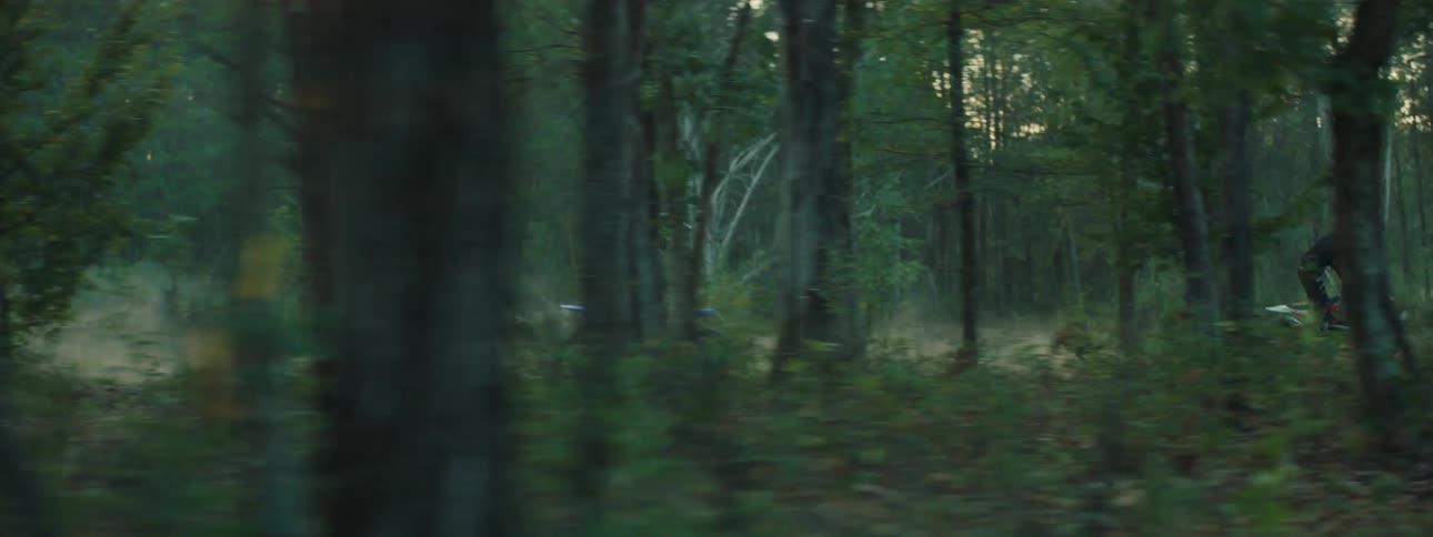 A motocross rider in dark gear speeds through a dense shadowy forest on a blue dirt bike the camera tracks their movement through the trees as they navigate the dusty trail and kick up a cloud of earth