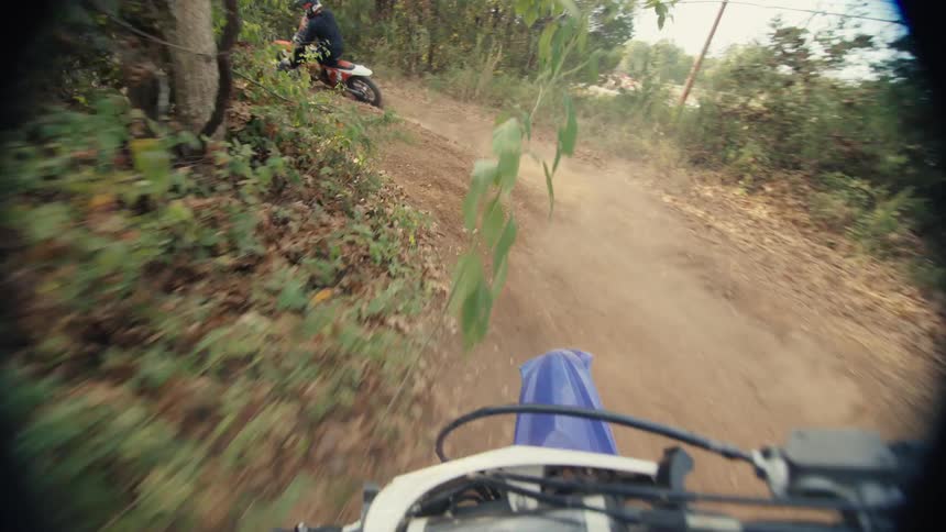 The video captures a firstperson perspective of a dirt bike rider chasing another cyclist through a narrow wooded trail the highspeed pursuit kicks up clouds of dust as they navigate sharp turns and uneven terrain under dappled sunlight