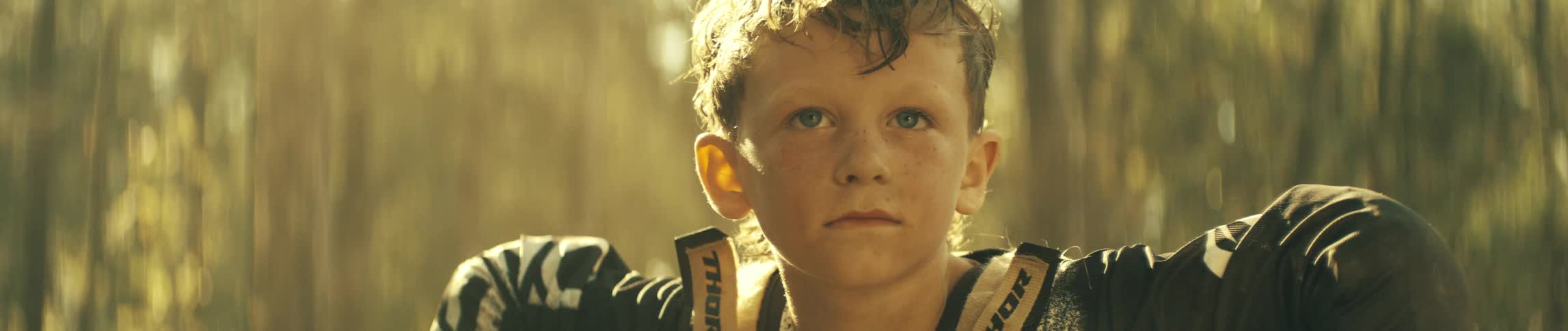 A young motocross rider with dirtspeckled cheeks stares ahead with quiet determination golden forest light glowing behind him