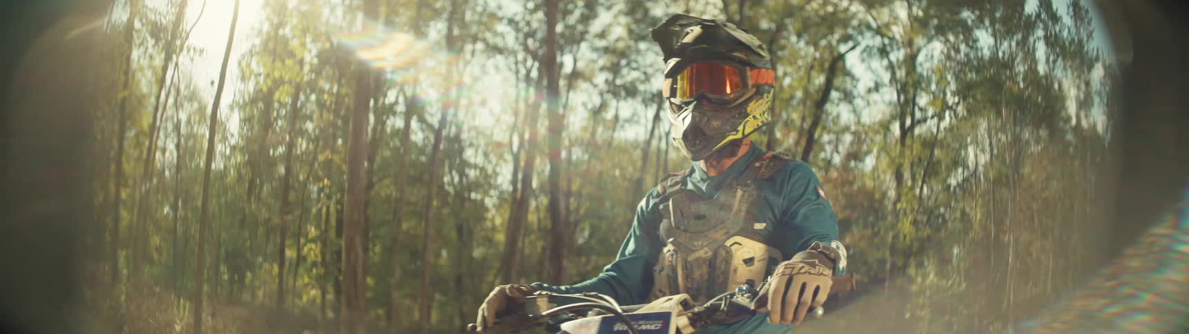 A motocross rider wearing full protective gear sits on a motorcycle in a sunny forest while looking toward the side as light filters through the dense trees behind him