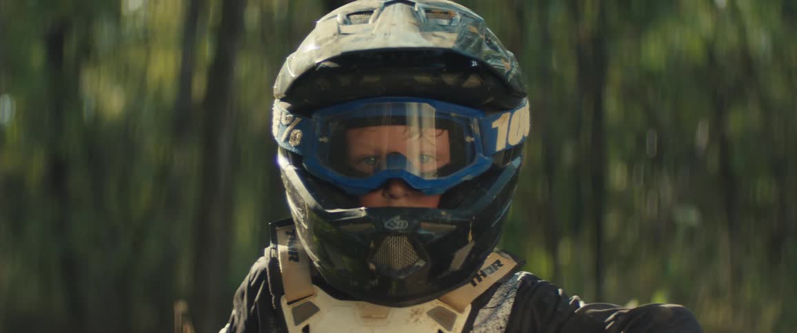 A young boy wearing a dirtspeckled motocross helmet and blue goggles stares intently forward taking a quiet breath against the softly blurred background of a sunlit forest