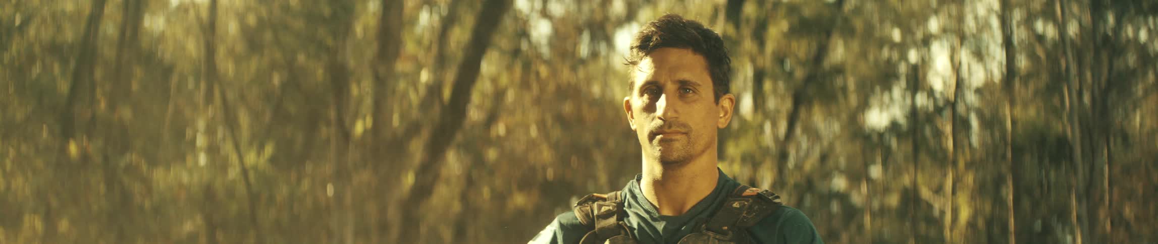 A man wearing riding gear stands in a forest bathed in warm golden sunlight looking ahead with a serious expression