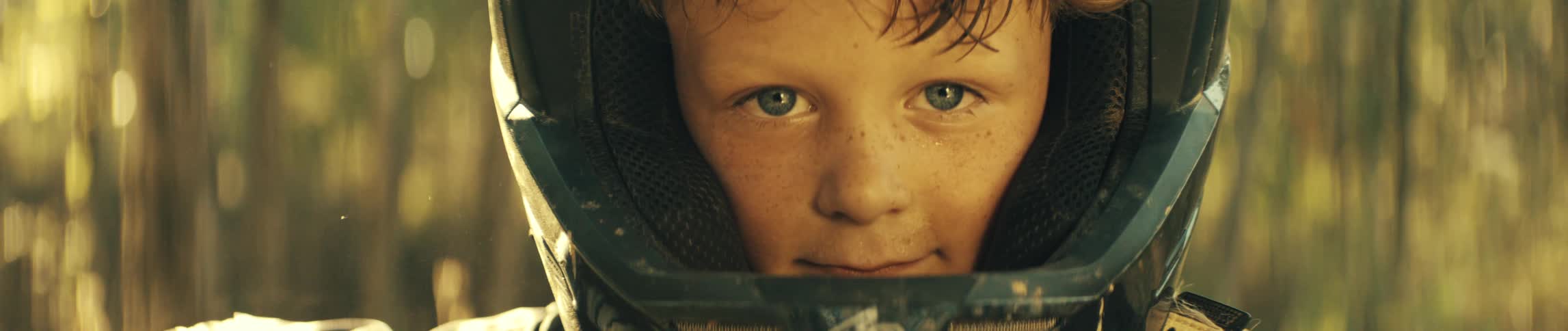 An extreme closeup captures the face of a young boy with blue eyes and freckles framed by a dark dirtspeckled helmethe looks directly into the camera with a subtle determined expression as sunlight highlights his features