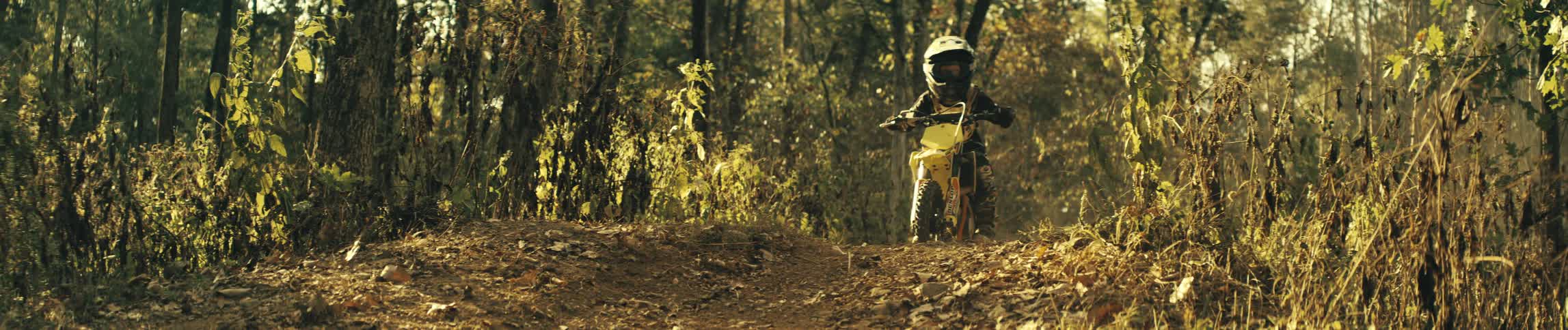 Boy rides a dirt bike on a forest dirt trail under sunlight among trees
