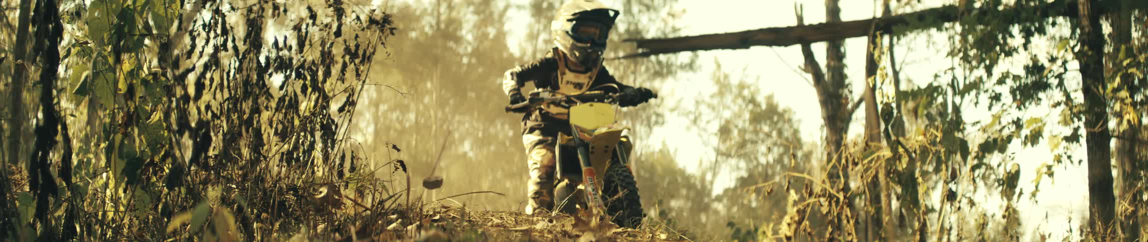 A rider wearing full protective gear and a helmet drives a yellow dirt bike over a dusty forest trail toward the camera as sunlight filters through the trees and kicks up dry leaves