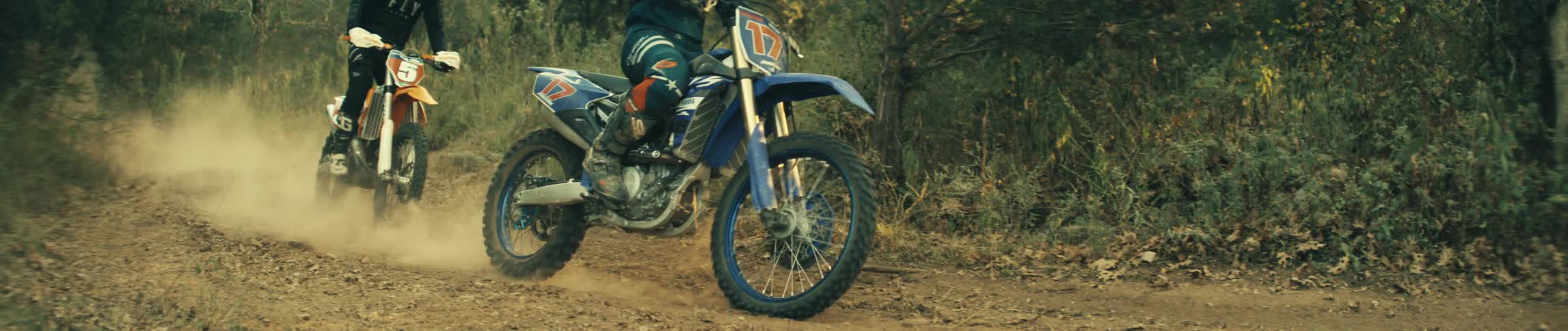 Two motorcyclists riding offroad bikes on a dirt trail with dust rising around them