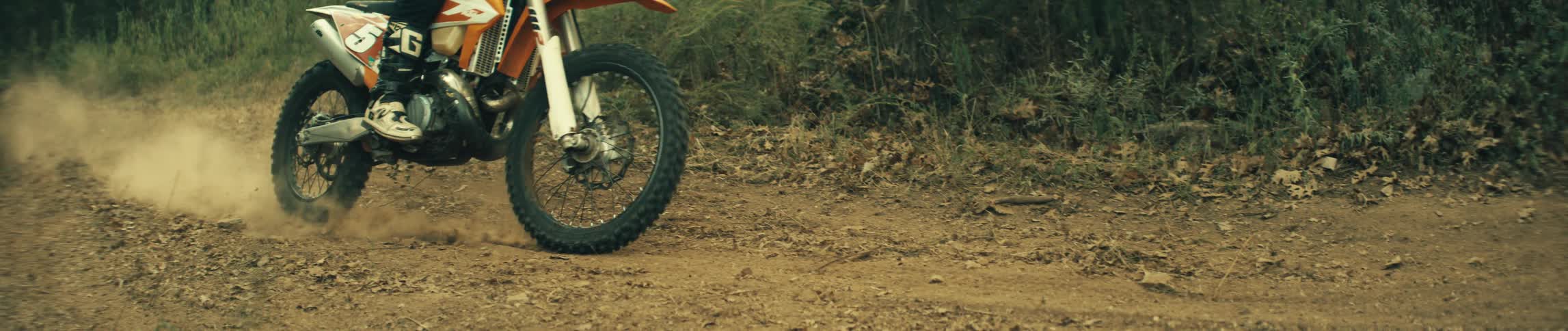 A close view of an orange and white motorcycle wheel as it turns and kicks up brown dust on a dirt path with green bushes in the background