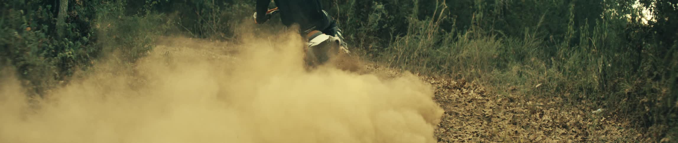 A dirt bike accelerates rapidly on a dry trail kicking up a massive thick cloud of brown dust from its rear tire the rider wearing full protective gear and a helmet speeds away into the distance as the dust hangs heavily in the air