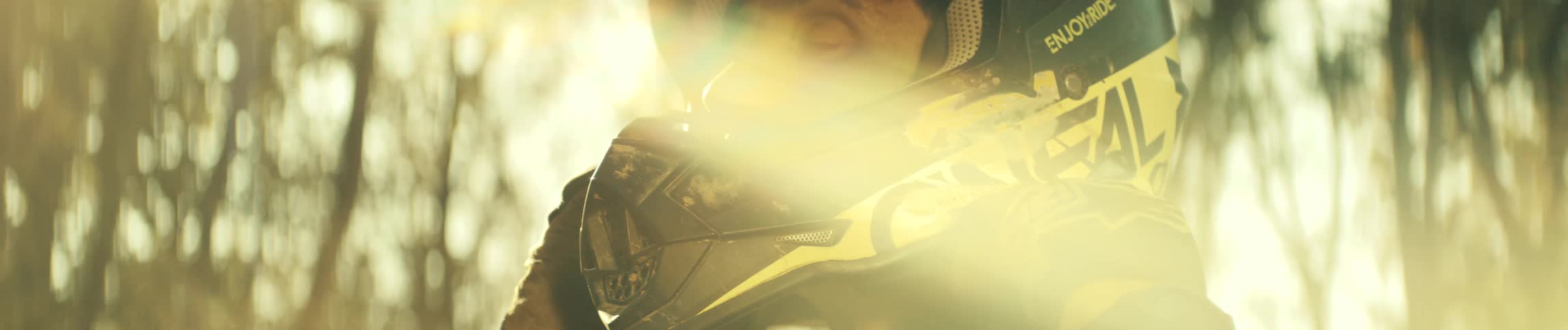 Man puts on a motocross helmet under sunlight in an outdoor setting with trees in the background