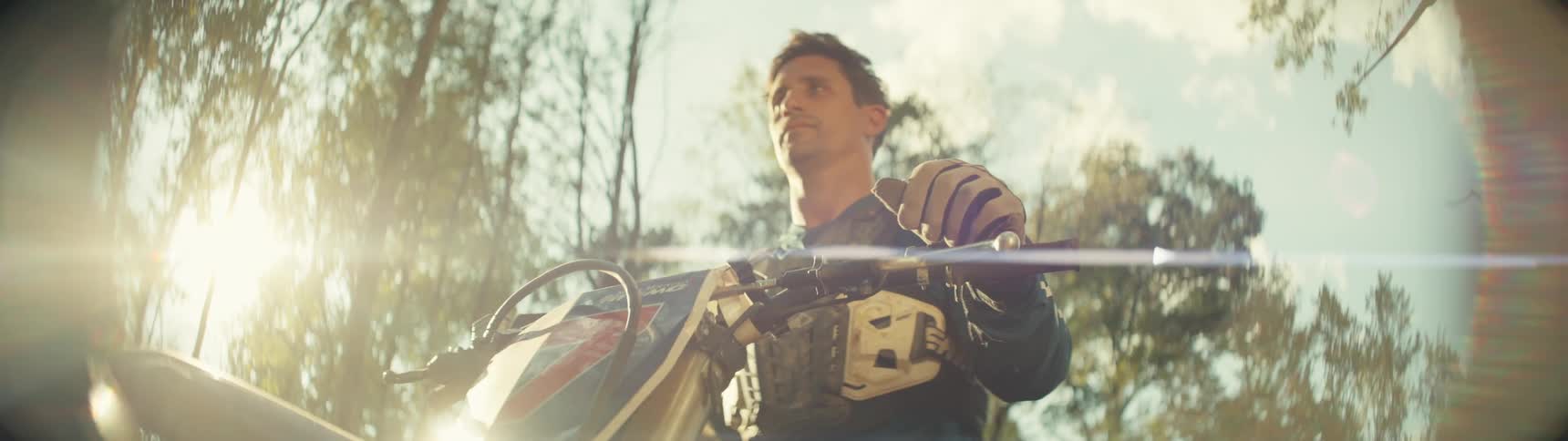 A motocross rider wearing a chest protector and gloves grips the handlebars of his dirt bike while looking into the distance under a bright sun filtered through tall trees in a forest