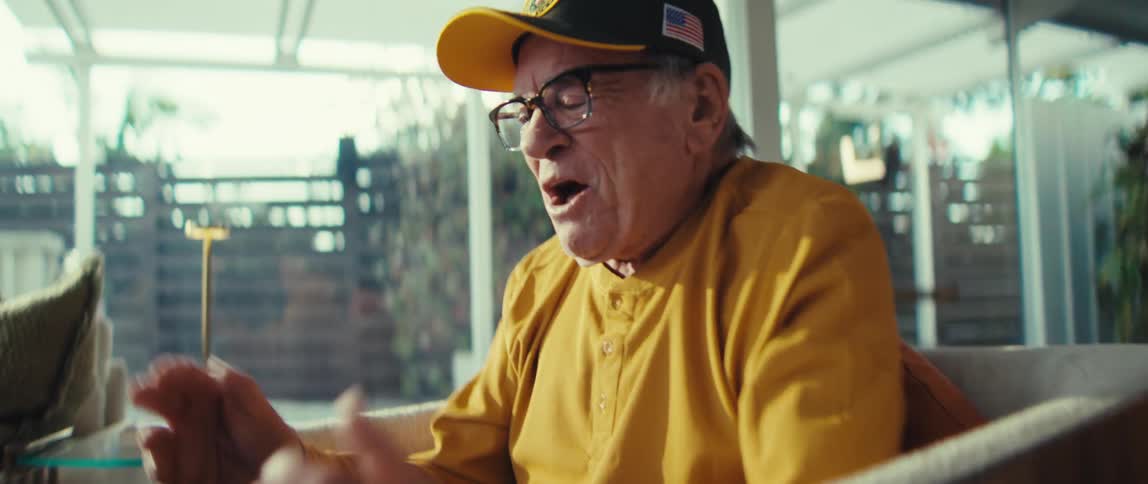 An elderly man in a yellow shirt and veteran cap laughs heartily looking at a table tablet while clapping his hands in a bright modern living room