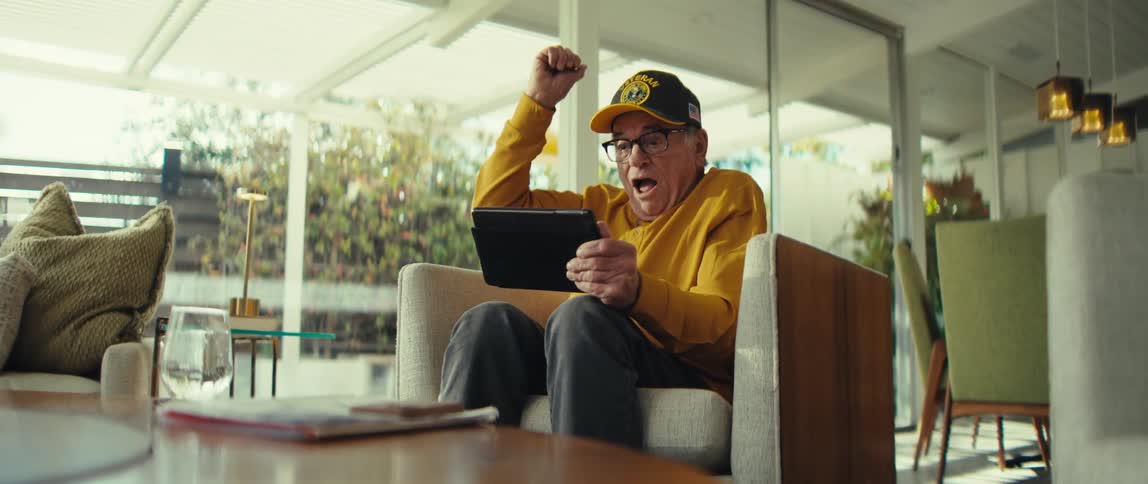 An older man wearing a yellow shirt and a veteran hat sits in an armchair while using a tablet and raises his fist in a celebrate motion with an excited expression in a bright living room