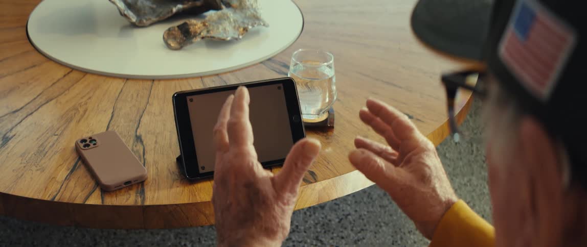 Elderly man wearing a cap and glasses sitting in front of a tablet computer with a replaceable screen over a table and cheering indoors during day time