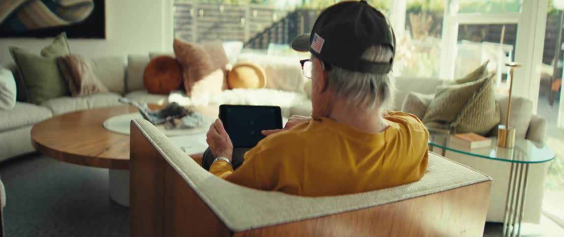 Man sitting in a wooden armchair wearing a cap and holding a tablet in a living room lit by natural light