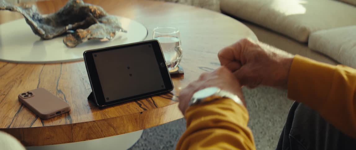 An older man wearing a yellow shirt and a watch sits at a table and pumps his fists in the air with excitement while looking at a tablet in a bright living room