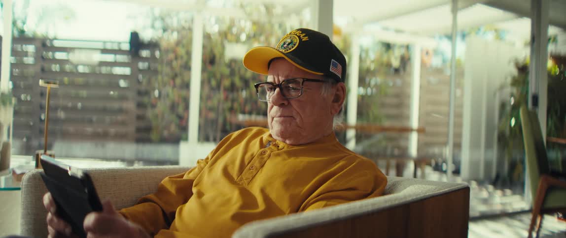 Elderly man wearing a cap and glasses sitting in an armchair while holding a tablet in a room with large windows in the background