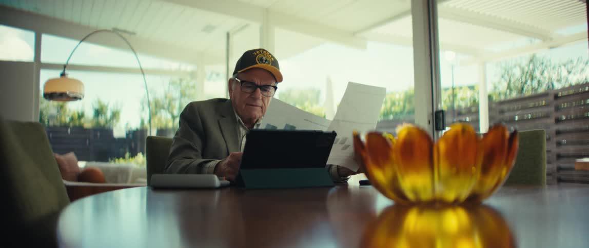 Elderly man wearing a cap and glasses sitting at a table analyzing reports in front of a tablet in a room with glass doors in the background