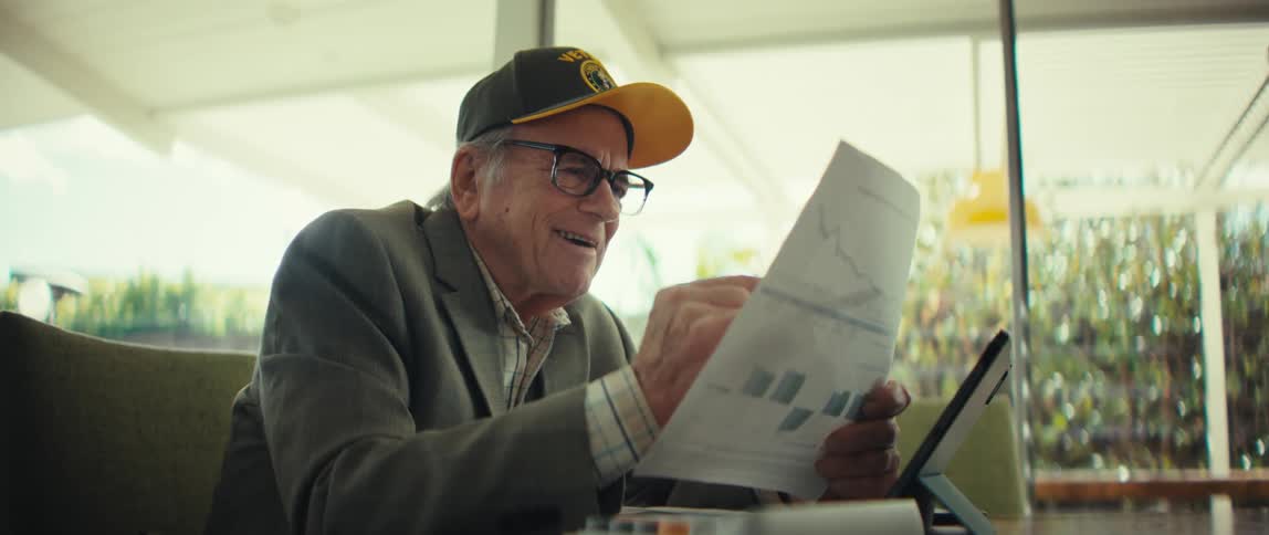 Elderly man with glasses wearing a blazer and a baseball cap sitting down and reading from a sheet of paper in front of a tablet computer while smiling indoors during day time