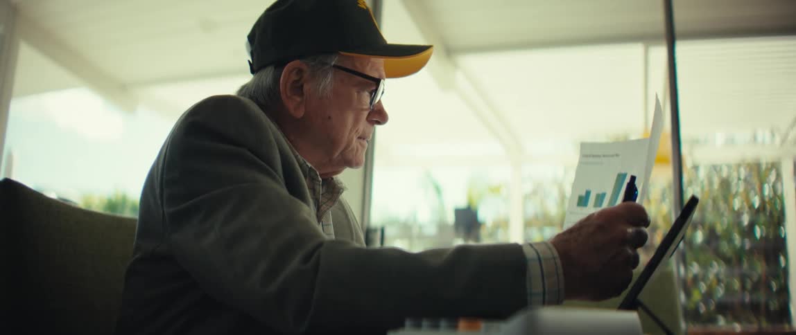 An elderly veteran wearing a black army cap and glasses sits at a table and looks through financial papers with blue and green bar graphs in a room with large windows