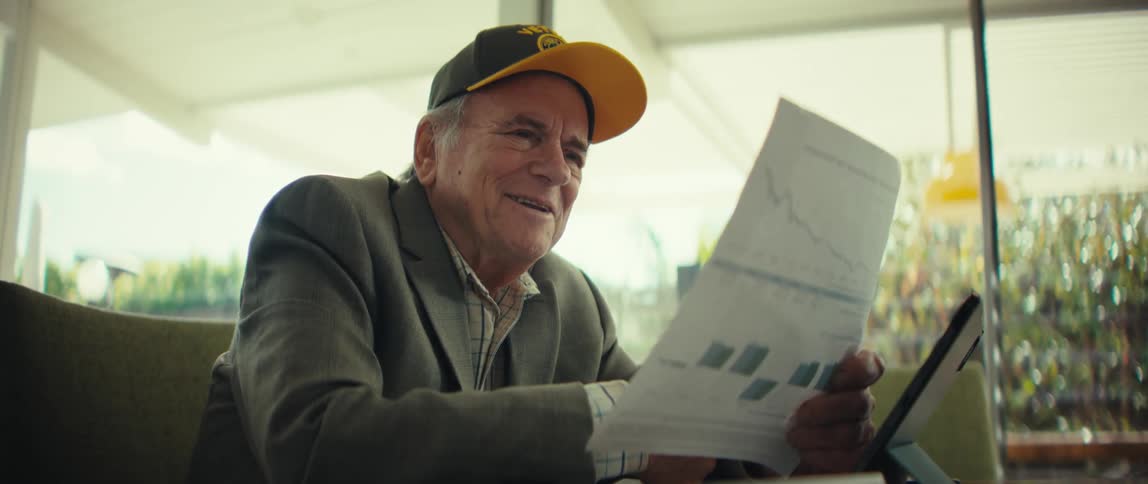 An older man wearing a baseball cap and glasses smiles and laughs while looking at a paper document with charts then removes his glasses and claps his hands